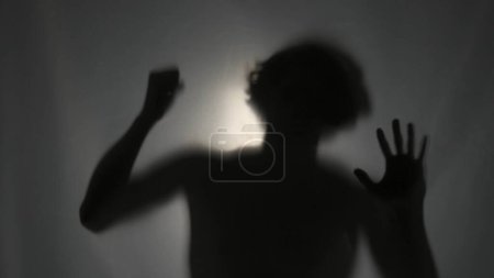 Téléchargez les photos : La silhouette sombre d'un homme derrière un rideau mat, illuminé par un faisceau de lumière. L'homme bat son poing, essayant de briser la barrière invisible. Le concept d'un fantôme - en image libre de droit