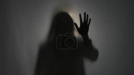 Photo for A dark silhouette of a woman behind a mat curtain, illuminated by a beam of light. The woman touches the curtain. The afterlife, the otherworld - Royalty Free Image