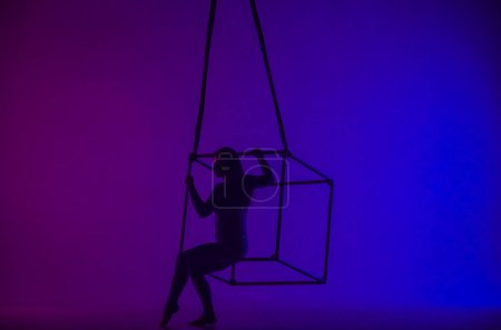 Photo for Silhouette of female aerial gymnast isolated on blue purple neon background. Female acrobat performing in the air on a cube, showing her flexibility - Royalty Free Image