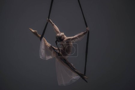 Foto de Chica gimnasta aérea demuestra estiramiento en cordel en trapecio acrobático. Atleta acrobático realiza colgando de una altura en un estudio oscuro - Imagen libre de derechos