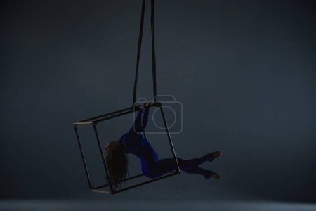 Téléchargez les photos : Silhouette de gymnaste aérien en justaucorps bleu avec strass sur fond studio sombre. acrobate féminin se produisant dans les airs sur un cube, exécutant des éléments de chorégraphie acrobatique - en image libre de droit