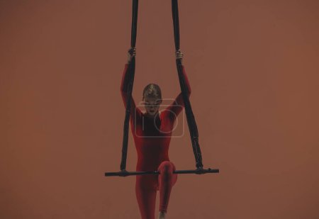 Foto de Gimnasta aérea femenina sobre fondo naranja del estudio. Chica artista de circo en mono rojo realiza elemento acrobático en el trapecio de aire, colgando en altura - Imagen libre de derechos