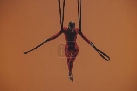 Téléchargez les photos : Femme gymnaste aérien sur fond orange du studio. Interprète de cirque fille en combinaison rouge exécutant un élément acrobatique avec des sangles suspendues en hauteur - en image libre de droit