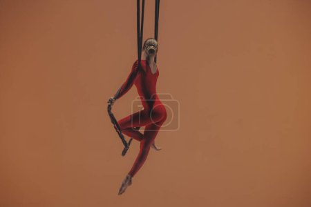 Photo for Female aerial gymnast on orange background of studio. Girl circus performer in red jumpsuit performing acrobatic element with straps hanging at height - Royalty Free Image