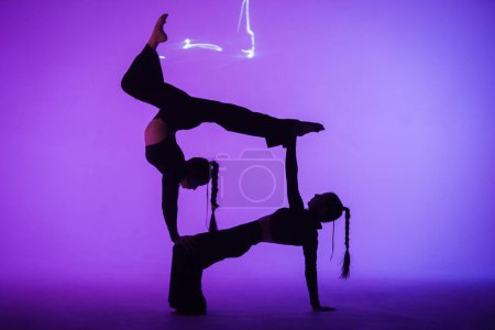 Téléchargez les photos : Chorégraphie moderne et acrobaties concept publicitaire créatif. Duo gymnastique féminin isolé sur fond de studio néon bleu laser. Filles danseuses montrant des éléments de gymnastique sur le sol. - en image libre de droit