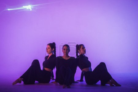 Foto de Trío de jóvenes acróbatas posando sentados en el suelo en el estudio con luz púrpura. Chicas gimnasta en trajes negros preparándose para la actuación - Imagen libre de derechos