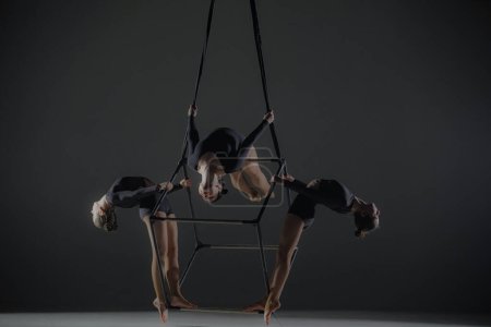 Foto de Trío de jóvenes acróbatas posando sobre un cubo suspendido a una altura. Los gimnastas aéreos actúan en estudio sobre fondo oscuro y demuestran flexibilidad y estiramiento. El concepto de espectáculo de circo - Imagen libre de derechos