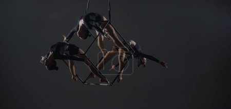 Téléchargez les photos : Trio de jeunes acrobates femelles posant sur un cube suspendu à une hauteur. Les gymnastes aériens se produisent en studio sur fond sombre et font preuve de flexibilité et d'étirement. Le concept de spectacle de cirque - en image libre de droit