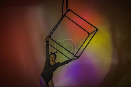 Foto de Gimnasta aérea en maillot negro sobre fondo multicolor de estudio. Acróbata hembra actuando sobre un cubo, interpretando elementos de coreografía acrobática - Imagen libre de derechos