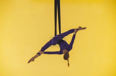 Téléchargez les photos : Chorégraphie moderne et acrobaties concept publicitaire créatif. Gymnaste femelle isolée sur fond jaune studio. Fille danseuse aérienne filant sur des sangles de gymnastique, montrant des éléments acrobatiques. - en image libre de droit