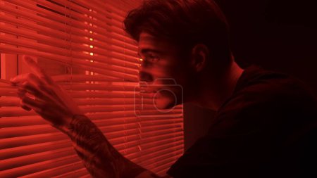 Photo for Silhouette abstract creative advertisement concept. Portrait of male in dark room. Handsome man near window, red neon light shines behind jalousie, guy opens blinds with hand peeks looks at window. - Royalty Free Image