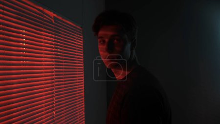 Téléchargez les photos : Silhouette abstraite concept publicitaire créatif. Portrait d'un homme dans une pièce sombre. Beau mec près de la fenêtre, néon rouge brille derrière jalousie, mec regarde la caméra, se retourne. - en image libre de droit