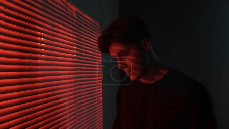 Photo for Silhouette abstract creative advertisement concept. Portrait of male in dark room. Handsome man near window, red neon light shines behind jalousie, guy looks down from camera, turns head. - Royalty Free Image