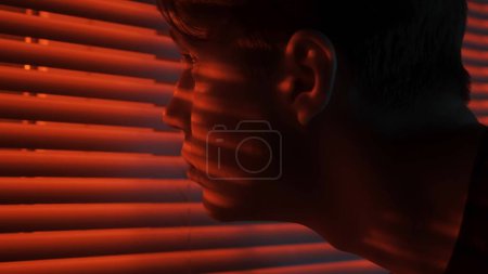 Téléchargez les photos : Silhouette abstraite concept publicitaire créatif. Portrait d'un homme dans une pièce sombre. Beau mec près de la fenêtre, néon rouge brille derrière jalousie, mec regarde dehors, plan de près extrême. - en image libre de droit