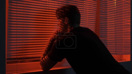 Téléchargez les photos : Silhouette abstraite concept publicitaire créatif. Portrait d'un homme dans une pièce sombre. bel homme près de fenêtre, néon rouge brille derrière jalousie, gars regarde dehors tenant la main sur le rebord de la fenêtre. - en image libre de droit