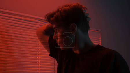 Photo for Silhouette abstract creative advertisement concept. Portrait of male in dark room. Handsome man near window, red neon light shines behind jalousie, guy looks outside and touches his hair. - Royalty Free Image