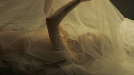 Téléchargez les photos : Vue latérale d'une jeune femme sur un lit, sous un chiffon transparent, gros plan. Une femme éclairée par une lumière chaude leva les bras, formant un tulle comme un auvent - en image libre de droit