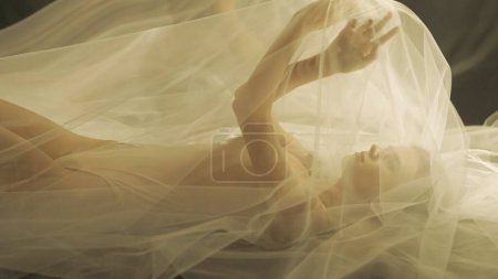 Photo for Side view of a young woman on a bed, under a transparent air cloth, close up. A woman illuminated by warm light raised her arms, forming a tulle like canopy - Royalty Free Image