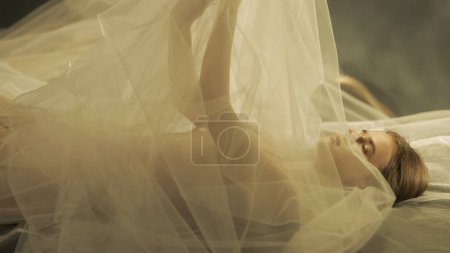 Photo for Side view of a young woman on a bed, under a transparent air cloth, close up. A woman illuminated by warm light raised her arms, forming a tulle like canopy - Royalty Free Image