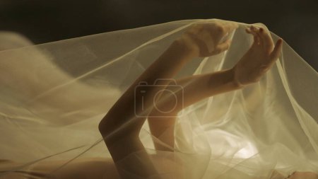 Foto de Manos de una mujer con tul blanco sobre un fondo negro de cerca. Un espejo refleja la silueta borrosa de una mujer cubierta con tela de red, iluminada por cálidos rayos de luz - Imagen libre de derechos