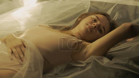 Téléchargez les photos : Portrait d'une femme naturelle couchée sur le dos sur un tissu transparent blanc au soleil en gros plan. Une femme en maillot de bain couleur chair regarde droit devant elle avec un regard pénétrant. Le concept de - en image libre de droit