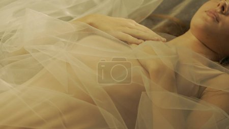 Téléchargez les photos : Mains de femme avec manucure nue, pliée sur la poitrine des femmes, gros plan. Femme couchée sur le dos couverte de tulle blanc, éclairée par des rayons de lumière chauds - en image libre de droit