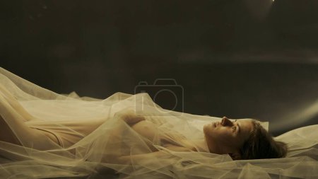 Photo for Side view of a woman lying motionless on her back with her arms folded across her chest near a mirror. The woman is covered up to her neck with a white transparent tulle cloth, illuminated by warm - Royalty Free Image