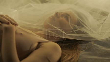 Photo for An attractive woman lying motionless on her back with her arms folded on her chest, close up. The womans face is covered with a white mesh cloth like a veil in a warm light - Royalty Free Image