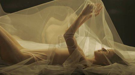 Téléchargez les photos : Vue latérale d'une jeune femme sur un lit, sous un chiffon transparent, gros plan. Une femme éclairée par la lumière chaude se reflète dans un miroir liquide - en image libre de droit