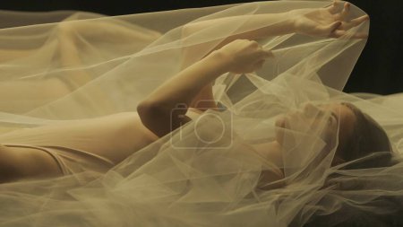 Téléchargez les photos : Vue latérale d'une jeune femme sur un lit, sous un chiffon transparent, gros plan. Une femme éclairée par la lumière chaude se reflète dans un miroir liquide - en image libre de droit