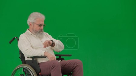 Téléchargez les photos : Pensionné vie quotidienne concept publicitaire créatif. Portrait d'homme âgé sur fond vert chroma key. Homme âgé assis dans un fauteuil roulant et regardant l'horloge - en image libre de droit