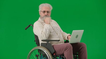 Photo for Pensioner everyday life creative advertisement concept. Portrait of aged man on chroma key green screen background. Senior man sitting in wheelchair and holding laptop on knees, watching video. - Royalty Free Image