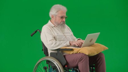 Photo for Pensioner everyday life creative advertisement concept. Portrait of aged man on chroma key green screen background. Senior man sitting in wheelchair and holding laptop on pillow, watching video. - Royalty Free Image