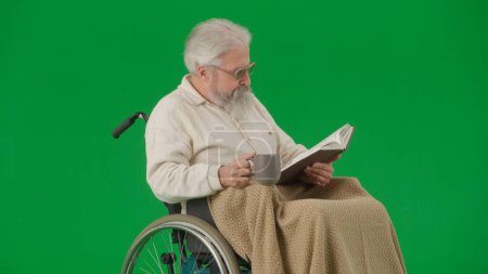 Photo for Pensioner everyday life creative advertisement concept. Portrait of aged man isolated on chroma key green screen background. Senior man in wheelchair covered by plaid reading book holding cup of tea. - Royalty Free Image