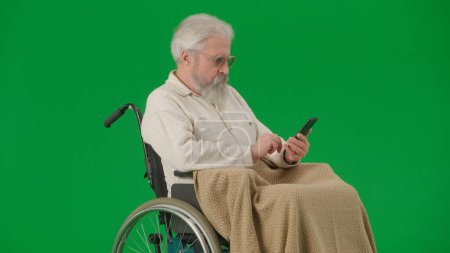 Téléchargez les photos : Pensionné vie quotidienne concept publicitaire créatif. Portrait d'un homme âgé isolé sur fond vert chromatique. Homme âgé en fauteuil roulant couvert par le balayage à carreaux sur smartphone. - en image libre de droit