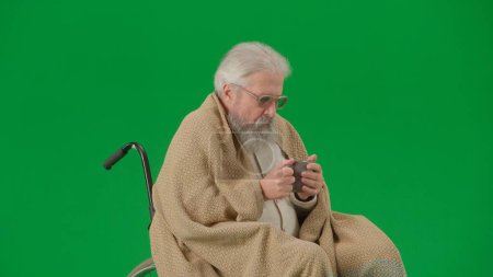 Photo for Pensioner everyday life creative advertisement concept. Portrait of aged man isolated on chroma key green screen background. Senior man in wheelchair covered by plaid drinking tea. - Royalty Free Image