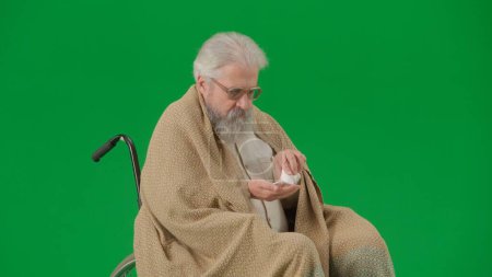 Photo for Pensioner everyday life creative advertisement concept. Portrait of aged man isolated on chroma key green screen background. Senior man in wheelchair covered by plaid taking pills from a bottle. - Royalty Free Image