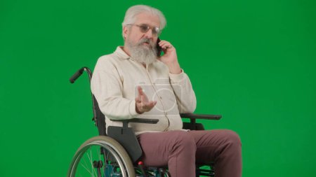 Photo for Pensioner everyday life creative advertisement concept. Portrait of disabled man isolated on chroma key green screen background. Senior man sitting in wheelchair talking on smartphone, full shot. - Royalty Free Image