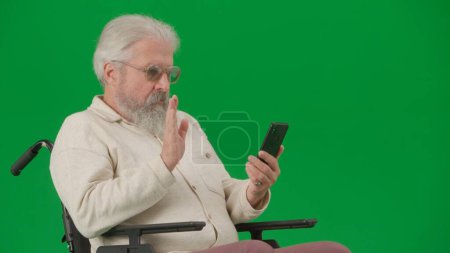 Photo for Pensioner caregiving and everyday life creative advertisement concept. Portrait of aged disabled man on chroma key green screen. Close up senior man in wheelchair talking video call on smartphone. - Royalty Free Image