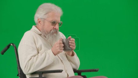 Photo for Pensioner everyday life creative advertisement concept. Portrait of disabled man isolated on chroma key green screen close up. Senior man sitting in wheelchair holding cup of tea and drinking. - Royalty Free Image