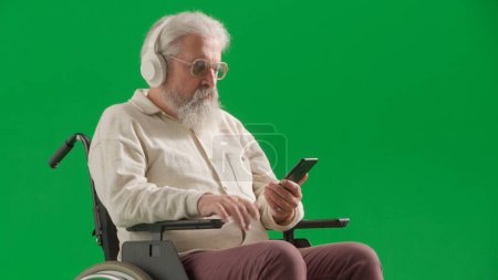 Téléchargez les photos : Pensionné vie quotidienne concept publicitaire créatif. Portrait d'un homme handicapé isolé sur écran vert chromatique de près. Homme âgé assis en fauteuil roulant portant un casque tenant un smartphone. - en image libre de droit