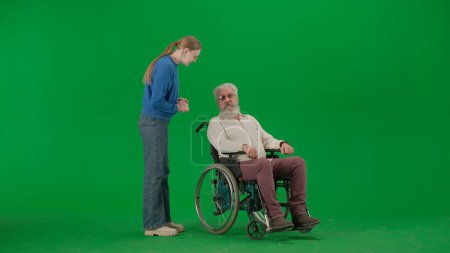 Photo for Pensioner everyday life creative advertisement concept. Portrait of aged man on chroma key green screen background. Young girl standing and talking to senior man granddad on a wheelchair. - Royalty Free Image