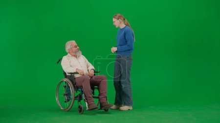 Photo for Pensioner everyday life creative advertisement concept. Portrait of aged man on chroma key green screen background. Young girl standing and chatting with senior man granddad on a wheelchair. - Royalty Free Image