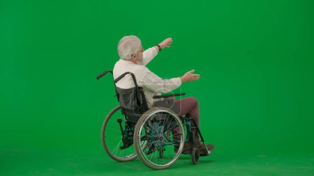 Téléchargez les photos : Pensionné vie quotidienne concept publicitaire créatif. Portrait d'homme âgé sur fond vert chroma key. Un aîné assis en fauteuil roulant montre des directions et des gestes avec les mains. - en image libre de droit