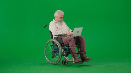 Foto de Pensionista vida cotidiana concepto de publicidad creativa. Retrato del hombre de edad en croma clave fondo de pantalla verde. Hombre mayor sentado en silla de ruedas y sosteniendo el ordenador portátil en las rodillas, viendo vídeo. - Imagen libre de derechos