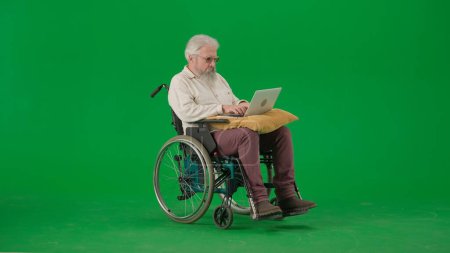 Téléchargez les photos : Pensionné vie quotidienne concept publicitaire créatif. Portrait d'homme âgé sur fond vert chroma key. Homme âgé assis en fauteuil roulant et tenant un ordinateur portable sur l'oreiller, regarder la vidéo. - en image libre de droit