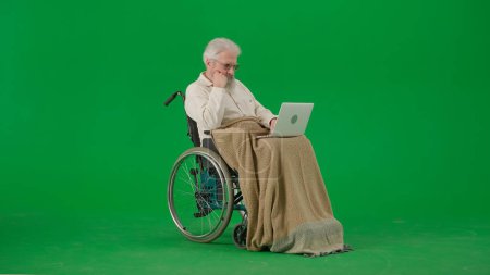 Photo for Pensioner everyday life creative advertisement concept. Portrait of aged man isolated on chroma key green screen background. Senior man in wheelchair covered by plaid watching video on laptop. - Royalty Free Image