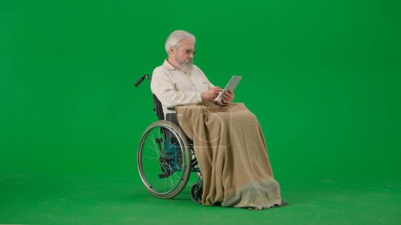 Photo for Pensioner everyday life creative advertisement concept. Portrait of aged man isolated on chroma key green screen background. Senior man in wheelchair covered by plaid swiping on tablet. - Royalty Free Image