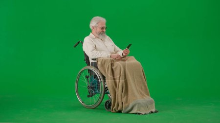Photo for Pensioner everyday life creative advertisement concept. Portrait of aged man isolated on chroma key green screen background. Senior man in wheelchair covered by plaid holding smartphone. - Royalty Free Image