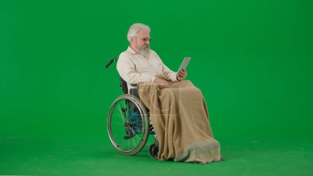 Photo for Pensioner everyday life creative advertisement concept. Portrait of aged man isolated on chroma key green screen background. Senior man in wheelchair covered by plaid holding tablet. - Royalty Free Image